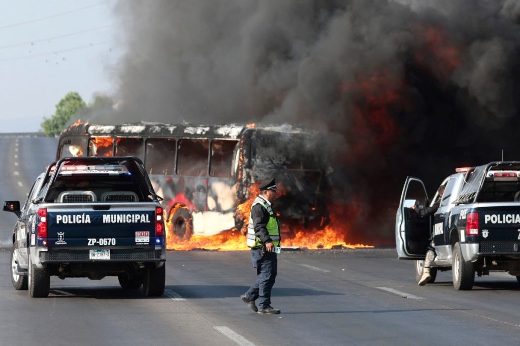 Camionetas de la policía municipal permanecen junto a un autobús en llamas, incendiado por grupos del crimen organizado en respuesta a un operativo en Jalisco para detener a un objetivo de seguridad de alta prioridad. /AFP