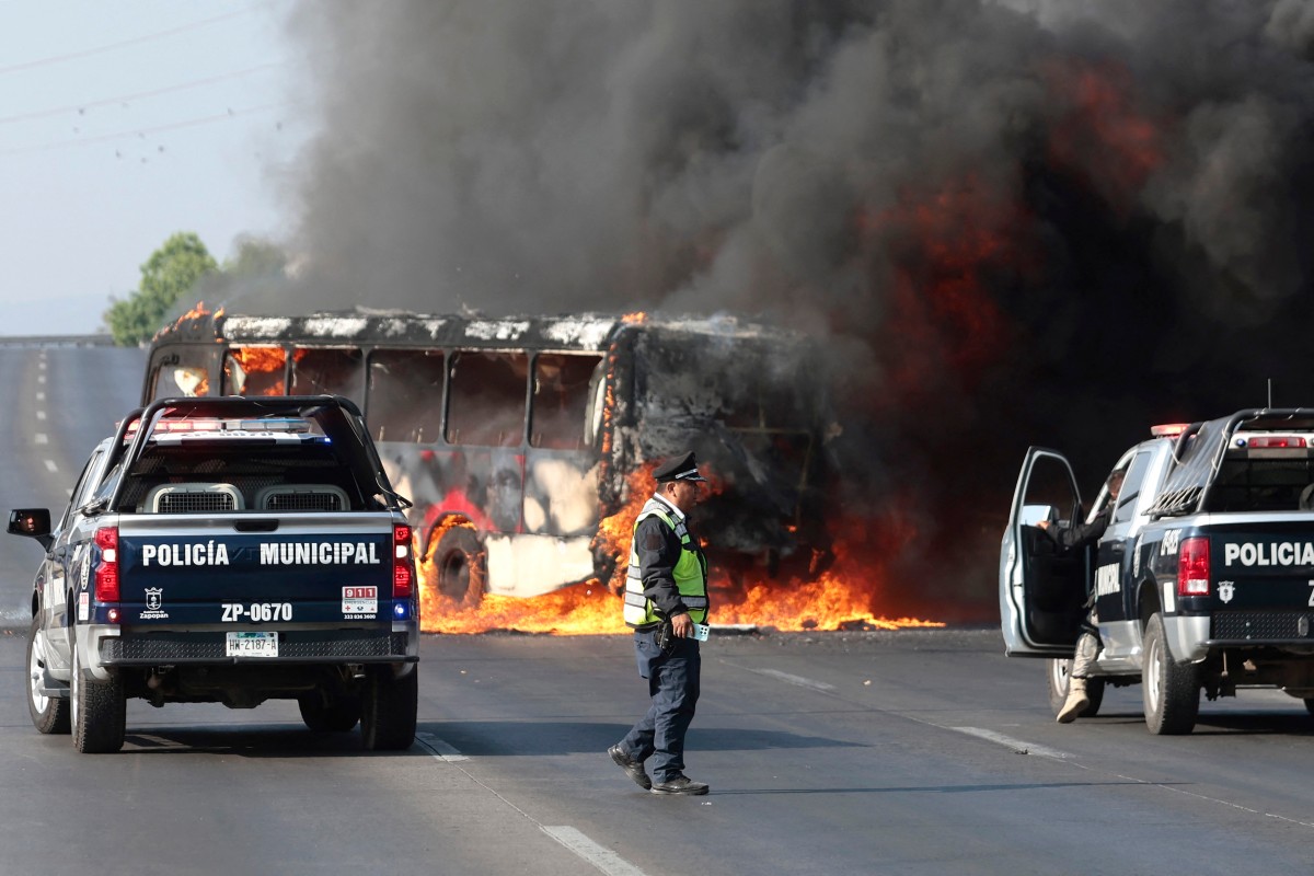 Camionetas de la policía municipal permanecen junto a un autobús en llamas, incendiado por grupos del crimen organizado en respuesta a un operativo en Jalisco para detener a un objetivo de seguridad de alta prioridad. /AFP